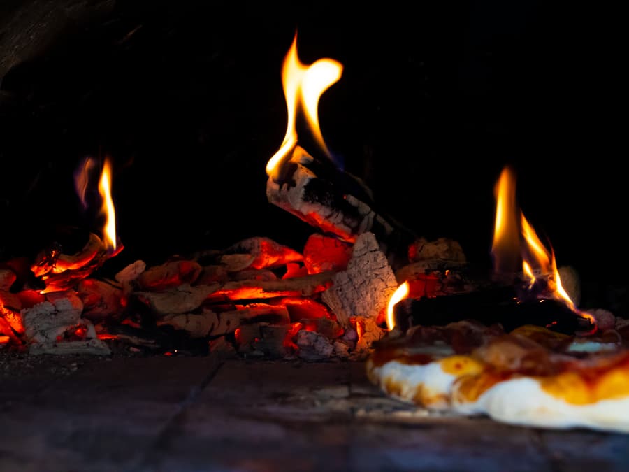 Braci e fiamme nel forno a legna, con una pizza in cottura sul piano