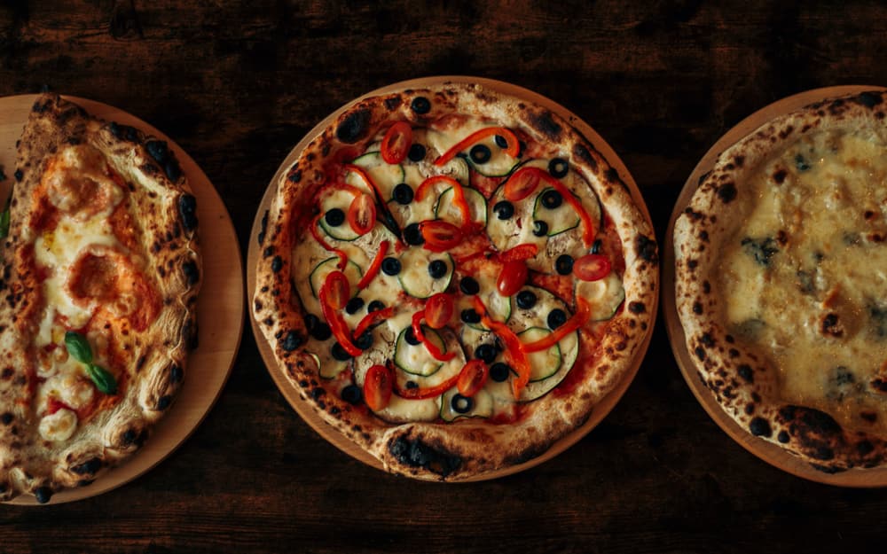 Tre pizze appena sfornate su assi di legno: una ripiena, una con verdure e olive, una bianca