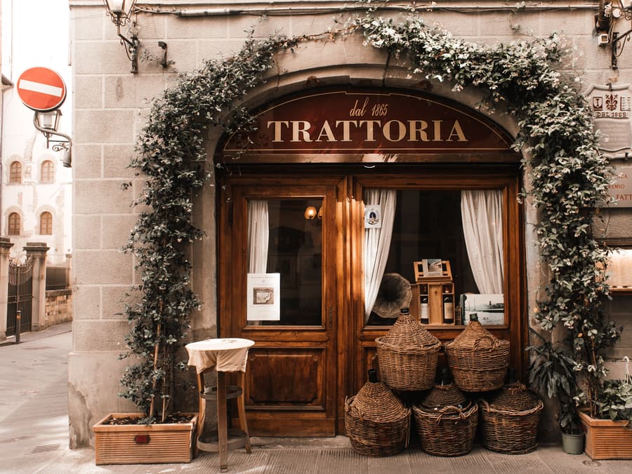 La facciata della trattoria con l’insegna in legno, l’edera intorno all’ingresso e le damigiane accanto alla porta