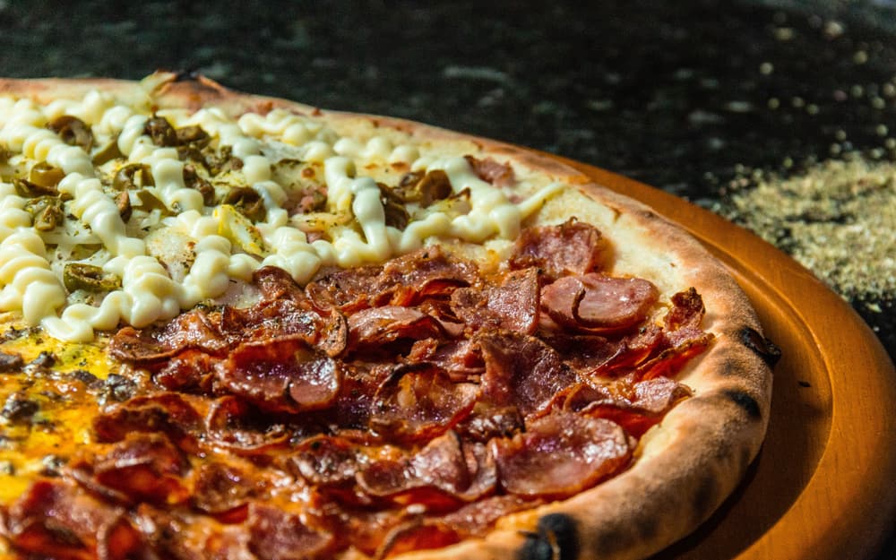 Pizza divisa a metà tra salumi e crema con olive verdi, servita su un piano di legno chiaro